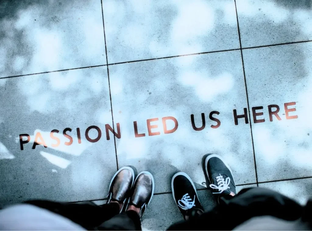 Overhead view of two pairs of shoes standing on a sidewalk with the words "PASSION LED US HERE" written across the pavement. The image symbolizes purpose-driven portfolio career design paths and is used in the context of portfolio career coaching by Andreia Fernandes at Seabrand. Photography by Ian Schneider (unsplash).